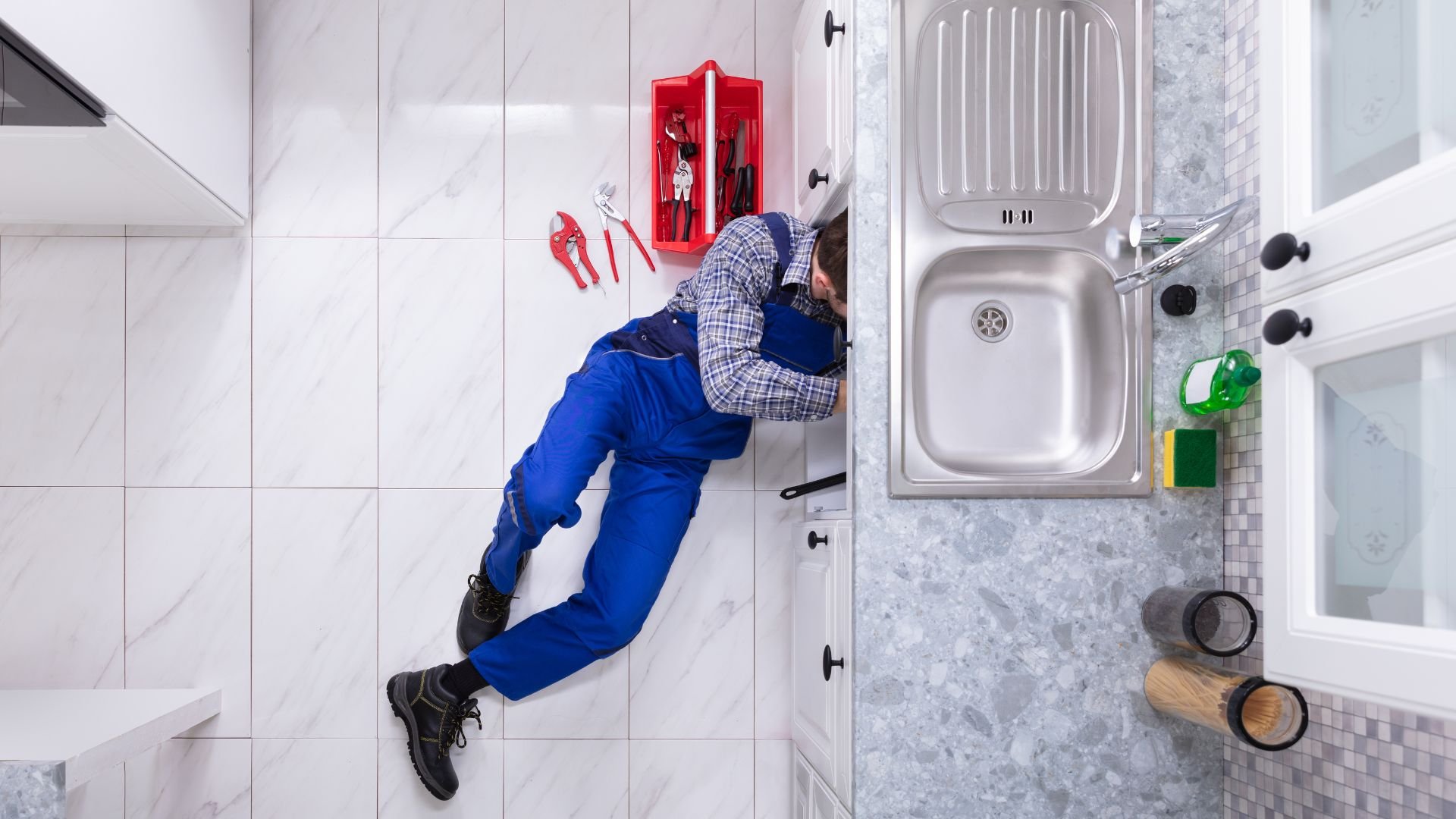 Plumber in blue overalls fixing sink pipes under kitchen cabinet