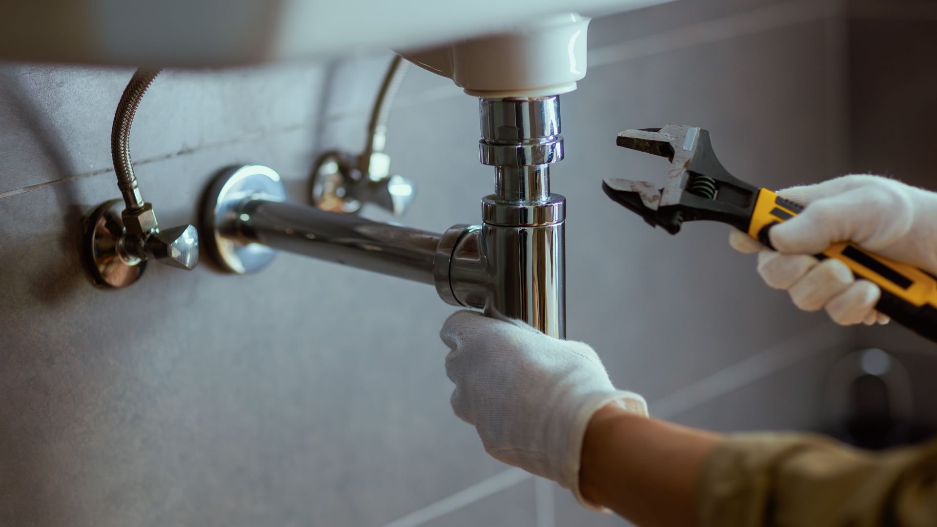 Hands in gloves fixing sink pipes with wrench under bathroom sink