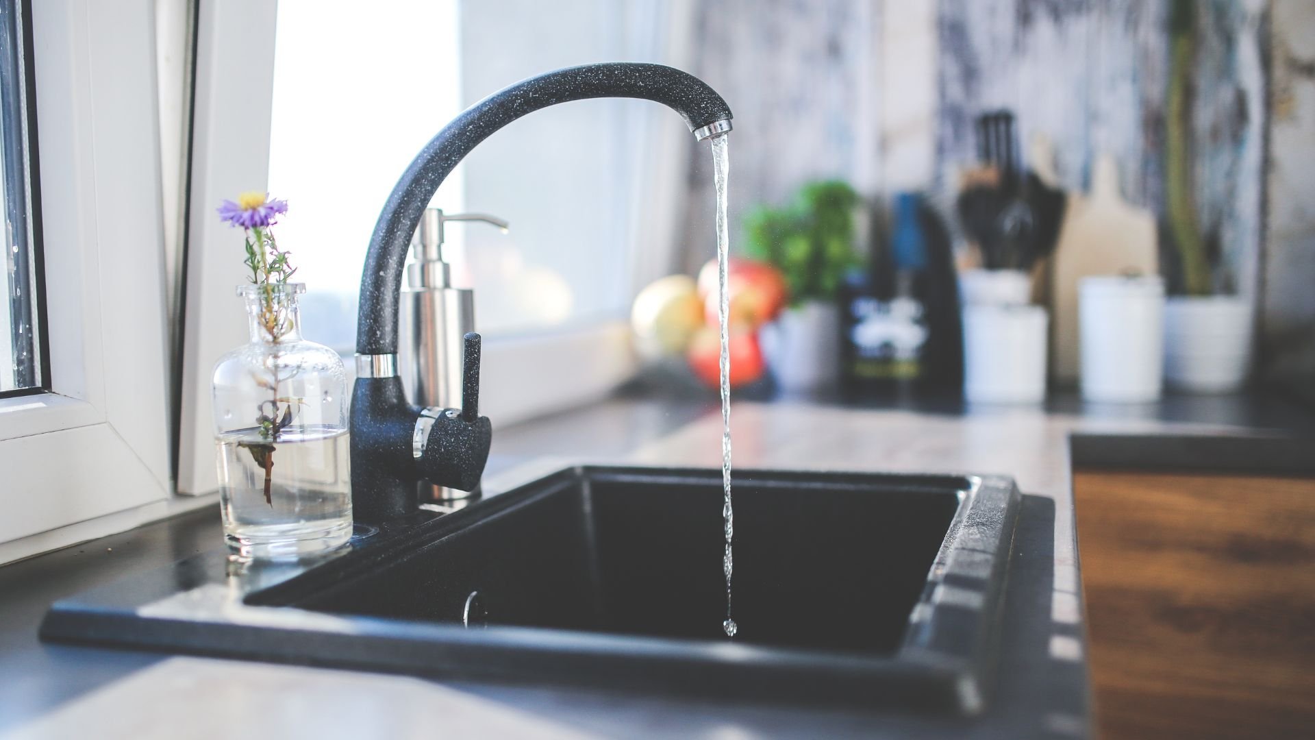 Water running from black kitchen faucet with small flower in glass vase